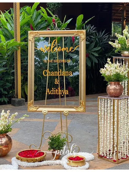 A modern and chic welcome board made of transparent acrylic with a gold frame. The setup is enhanced by copper pots with tuberoses and stands draped in floral strings.