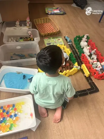 A city is being built, one sentence at a time. This setup invites a child to explore different sensory bins, from water beads to sand, creating a rich context for language to flourish naturally through play.