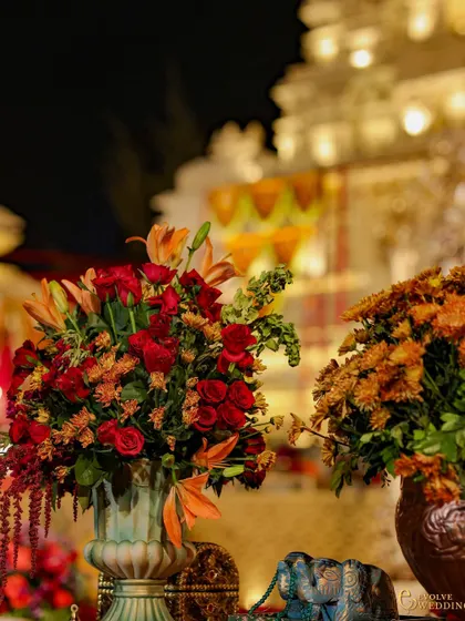 A detail shot of the vibrant floral arrangements, with the grand mandap softly blurred in the background. The rich reds and oranges of the flowers echo the warmth and opulence of the overall theme.