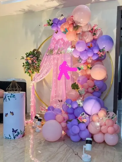 A whimsical butterfly-themed decor for a bridal shower or bachelorette party. The golden ring is draped with pink fabric and surrounded by a pastel balloon garland in shades of pink, purple, and peach, adorned with butterflies.