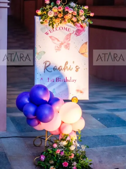 A beautiful welcome sign for Raahi's first birthday, decorated with a butterfly theme. The sign is surrounded by flowers and balloons, creating an elegant and inviting entrance for guests.
