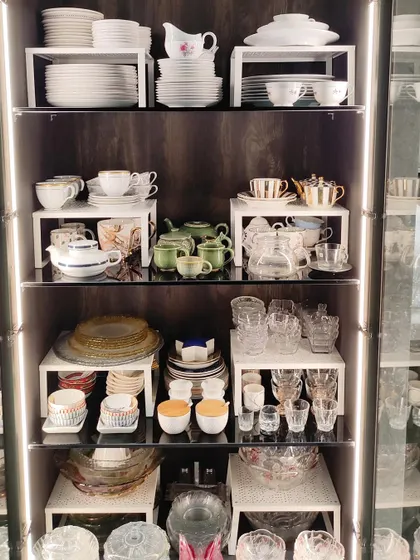 This beautifully lit crockery unit showcases a variety of tea sets and glassware. By using risers and grouping sets together, the collection is not only organized and safe but also visually stunning.