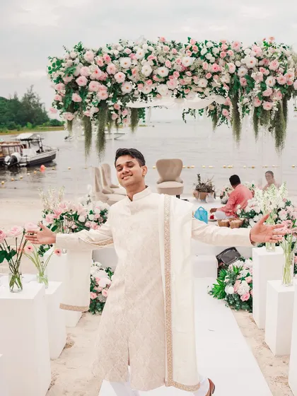 The groom, full of joy and excitement, stands at the floral mandap on a beach in Thailand, ready to welcome his bride. This candid shot perfectly captures the happy and relaxed vibe of a destination wedding.