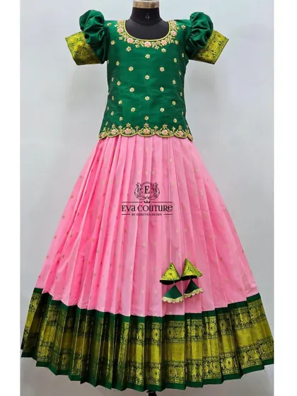 A classic pattu pavadai design with a pink skirt and a contrasting green blouse. The blouse has simple gold butti work, and the skirt features a traditional green and gold Kanchi border.
