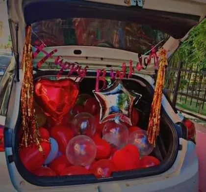 A romantic car boot surprise. The trunk is filled with red heart-shaped balloons and a 'Happy Birthday' banner, perfect for a celebration on the move.