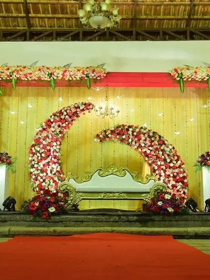 A vibrant wedding reception stage with a red and gold theme. The dual crescent floral arches create a unique focal point, perfect for the happy couple.