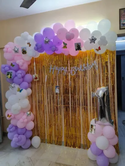 A first birthday decoration with a gold shimmer curtain and a pastel balloon arch. The arch is personalized with photos of the birthday girl.