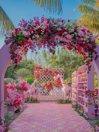 A whimsical and colorful entrance arch made of pink flowers, perfect for a daytime event. The path is flanked by playful giraffe statues, adding a unique and fun element to the decor.