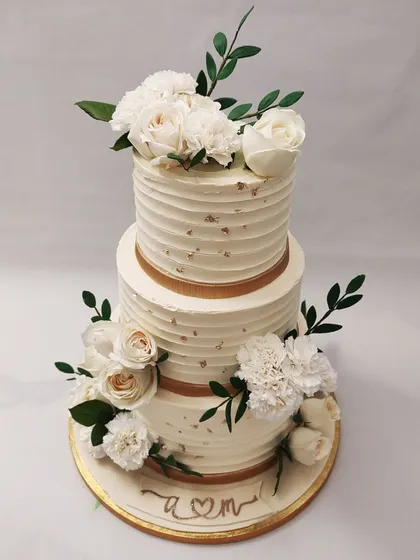 We design the picture of wedding day elegance with this royal white floral cake. Two tiers of textured buttercream are wrapped in gold ribbon and adorned with bouquets of fresh white roses and carnations, creating a timeless and sophisticated wedding centerpiece.