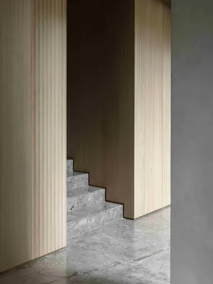 The clean lines of the marble staircase and the warm texture of the wood-paneled walls create a serene and sophisticated passage between floors. The design emphasizes material honesty and minimalist elegance.