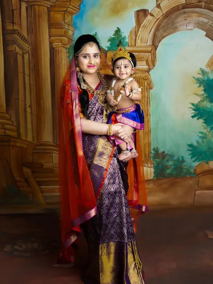 A beautiful portrait of a mother holding her baby, both dressed in traditional attire for a Janmashtami photoshoot. The love and pride in her eyes are unmistakable.