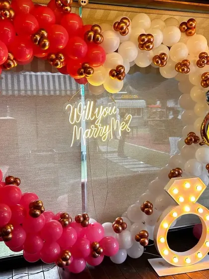 The perfect setup for a romantic proposal. This heart-shaped balloon arch in red and white features a 'Will you Marry Me?' neon sign and a light-up diamond ring prop.