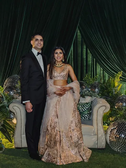 A stylish couple poses against a modern reception backdrop of emerald green velvet, lush ferns, and scattered disco balls for a touch of glamour.