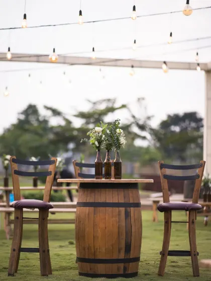 A rustic barrel table provides a charming spot for conversation. Paired with high chairs and simple bottle vases, it adds a touch of countryside flair to an outdoor Mehendi or cocktail event.