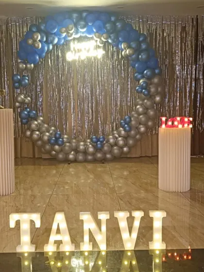A modern birthday setup with a silver tinsel backdrop, a blue and silver balloon ring, and illuminated letters spelling out the name "TANVI".