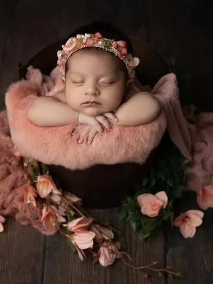 This baby girl looks like a sleeping fairy, nestled in a bucket with soft pink fabrics and surrounded by delicate flowers on a dark wood floor.