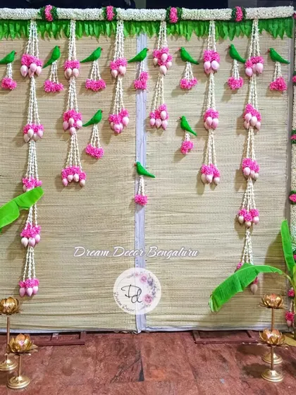 A classic and elegant baby shower backdrop. This design uses a traditional woven mat (chatai), jasmine and tuberose garlands, banana saplings, and brass lamps for an authentic South Indian feel.