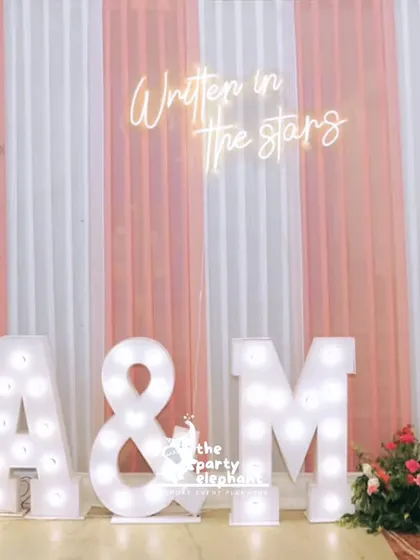 A personalized photo corner at the wedding reception, featuring large marquee letters of the couple's initials, a neon 'Written in the stars' sign, and beautiful floral arrangements.