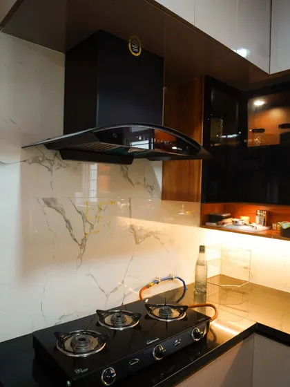 A close-up of a modern kitchen featuring a sleek black chimney against a white marble-veined backsplash. The under-cabinet lighting illuminates the granite countertop, highlighting the clean lines of the gas hob and the overall minimalist design.