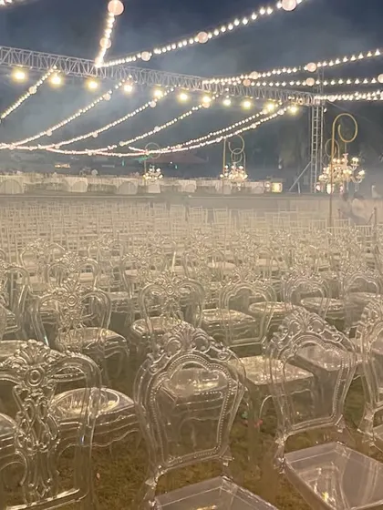 Preparing for a grand reception. Rows of elegant transparent ghost chairs are set up under a canopy of string lights and chandeliers, awaiting the arrival of guests for a magical evening.