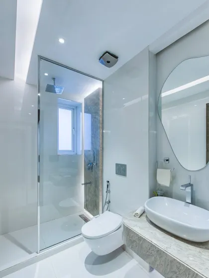 A bright and clean bathroom design with white marble walls and a frameless glass shower enclosure. The organic-shaped mirror adds a soft, contemporary touch to the minimalist space.