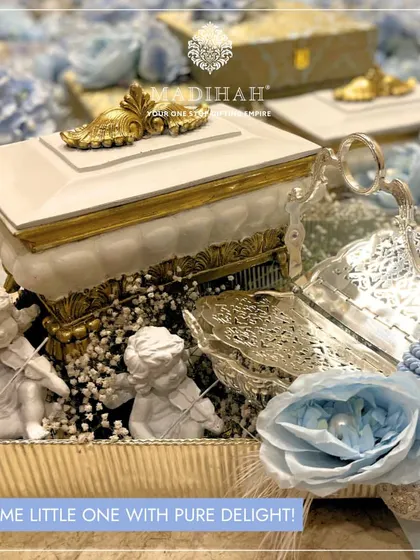 Welcome a little one with this exquisite hamper featuring cherubic angel figurines and soft blue floral arrangements. The design combines classic white and gold accents with silver-plated serveware, making it a perfect luxury gift for a baby boy's arrival.