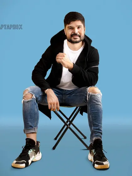 A seated portrait of a male model in a casual, layered outfit. The solid blue background makes the subject pop, and the direct-to-camera pose feels engaging and confident.