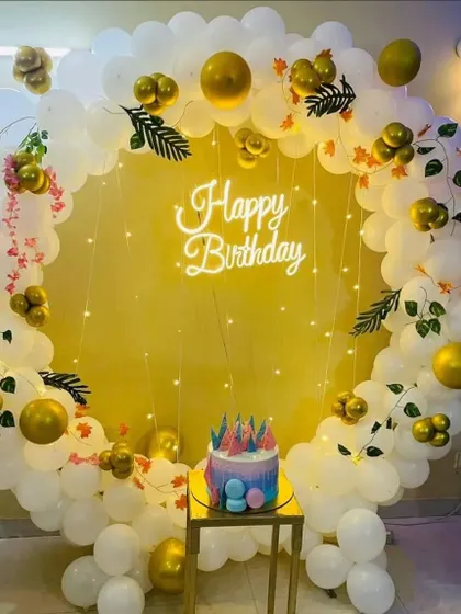 A beautiful birthday decoration with a circular balloon frame made of white and gold balloons. The setup includes a neon sign and is accented with artificial leaves for a touch of green.
