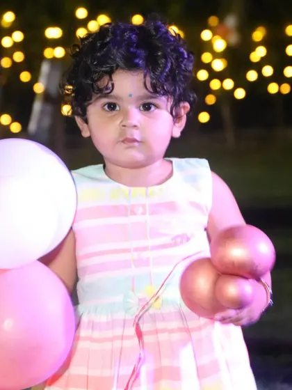 A close-up portrait of a toddler with curly hair holding balloons at her party. We specialize in kids' photography, capturing their innocent and curious expressions.