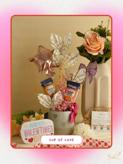 The 'Cup of Love' hamper is a compact and cute arrangement in a mug. It includes energy bars, a small balloon, and decorative elements, making it a perfect small token of affection.