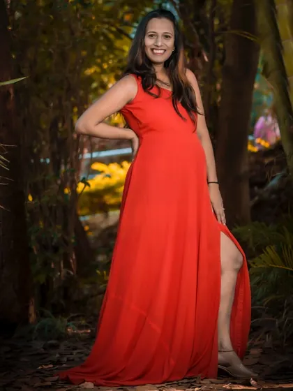 A confident and glamorous solo portrait in a striking red gown. The pose highlights both the baby bump and the elegance of the dress, set against a beautiful, warmly lit natural background.