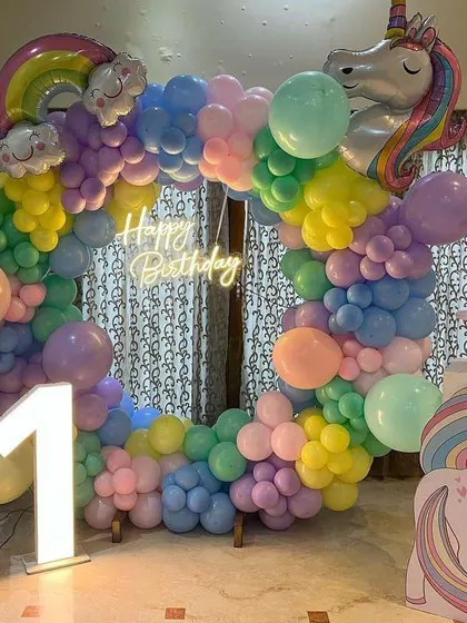 A circular rainbow balloon arch with a unicorn theme for an indoor celebration. The 'Happy Birthday' neon sign and light-up number '1' complete this magical setup.