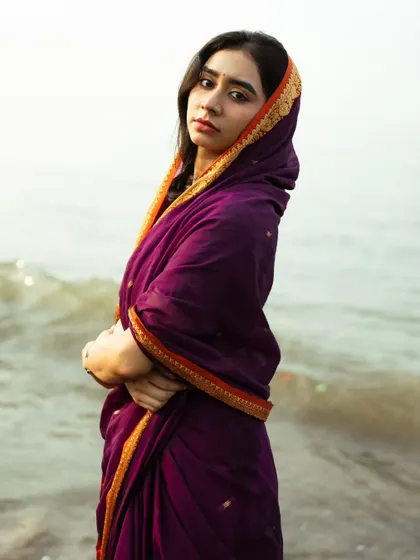 The deep purple of the saree against the muted tones of the sea creates a rich, moody, and almost painterly portrait.
