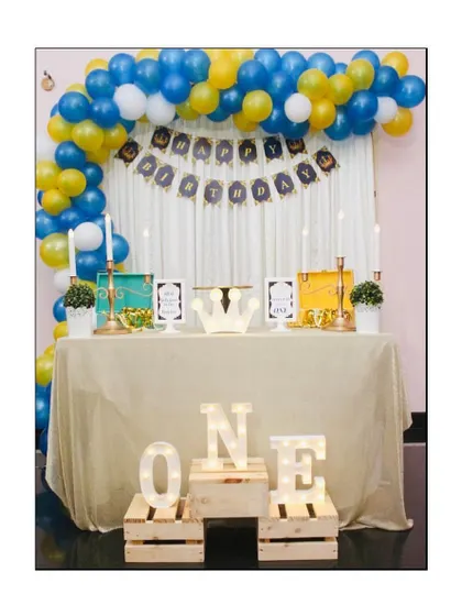 A royal prince theme for a first birthday party. The decor includes a blue, yellow, and white balloon arch, a personalized birthday banner, and light up 'ONE' letters on rustic wooden crates.