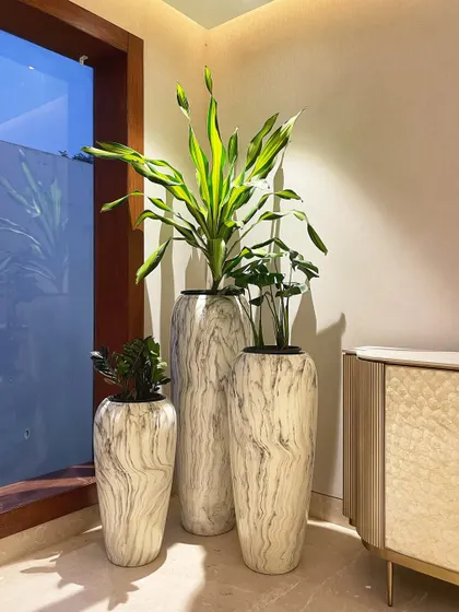 We love creating unique corner clusters. This trio of tall, marble-finish planters houses a Dracaena and other indoor plants, transforming an empty corner by the window into a sophisticated design feature.