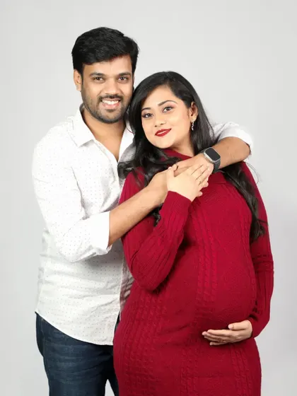 A classic and happy couple portrait. The mom-to-be is glowing in her red sweater dress while her partner holds her close, both smiling brightly for the camera.