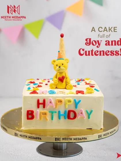 A cake full of joy and cuteness! This square birthday cake is decorated with colorful letters and a sweet yellow teddy bear wearing a party hat.
