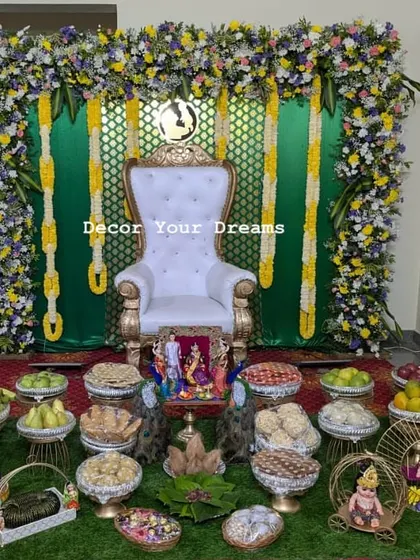 This home-based Seemantham setup features a green backdrop with floral hangings and a white throne. The floor is covered with artificial grass and a complete set of decorated offering plates.