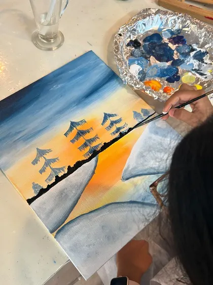 A close-up of a forest painting in progress. The artist is carefully adding the trees against the sunset sky.