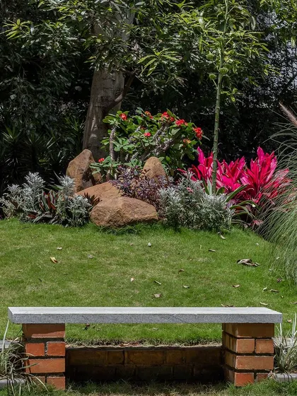 A simple bench made of a stone slab and brick supports is placed within a rock garden. The backdrop of boulders and colorful plants creates a natural and peaceful spot to sit and enjoy the surroundings.