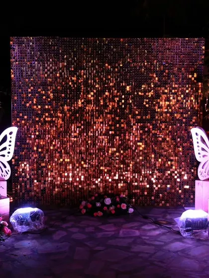 A dramatic rose-gold sequin wall set up outdoors for a marriage event, flanked by elegant white butterfly props for a touch of whimsy.