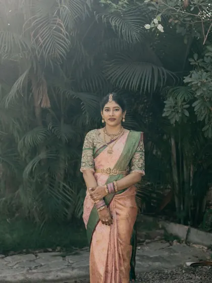 A full-length portrait of the bride. Her makeup and hair are styled to create a cohesive and elegant look that complements her traditional pink and green silk saree.