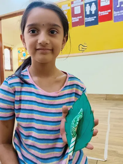 A young girl holds up her finished moving caterpillar craft. She is proud of her creation, which she made using paper, a stick, and some string.