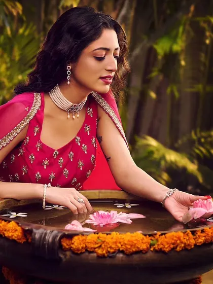 A serene moment from a festive jewellery campaign. The model, dressed in a vibrant pink outfit, interacts with lotus flowers in an urli, creating a peaceful and elegant scene that highlights the pearl choker and earrings.