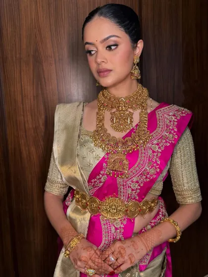 A regal bridal look featuring a pink and gold Kanchipuram saree. The drape is styled to showcase the grand temple jewelry and the intricate work on the saree border.