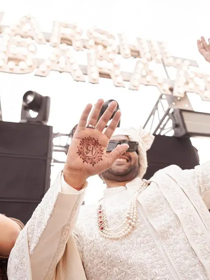 The groom shows off his mehendi, with the bride's initial creatively designed on his palm. This fun, candid shot captures a modern tradition and the festive spirit of the wedding.