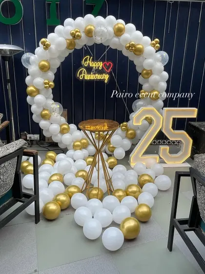 An elegant 25th-anniversary party backdrop. This features a full circle balloon arch in a sophisticated white and gold color scheme, a "Happy Anniversary" neon sign, and large "25" marquee numbers.