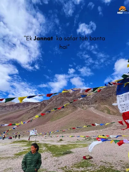 A traveler stands amidst prayer flags with the vast Spiti landscape behind her. A trip to Chandratal is truly a "jannat ka safar" (a heavenly journey).