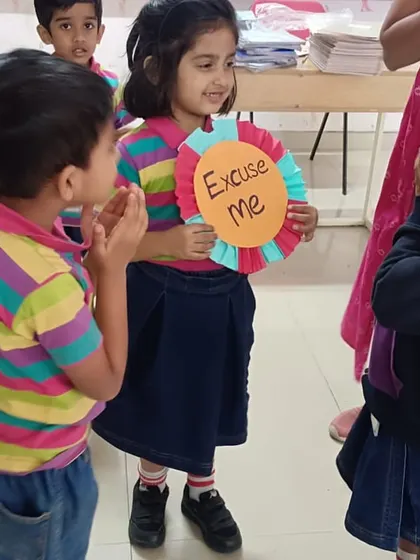 A student holds up an 'Excuse Me' sign while another listens attentively. Our interactive activities make learning social etiquette a memorable experience.