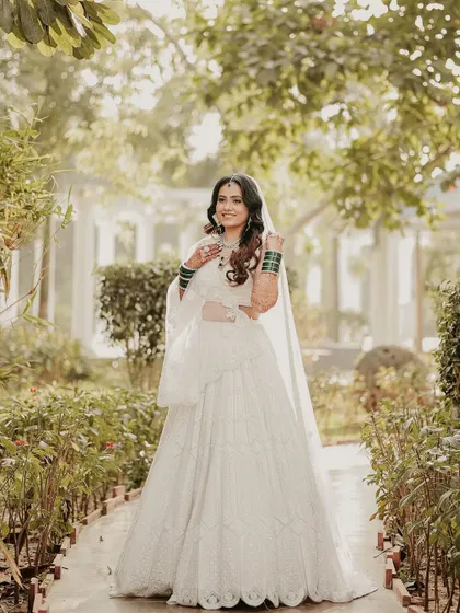 A full-length shot of the bride in a beautiful garden path. Her all-white lehenga stands out beautifully against the lush greenery, creating a fresh and elegant bridal portrait.